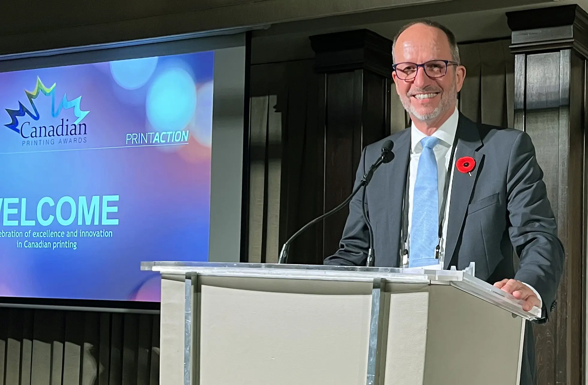 Ken Freek standing at the podium at the Canadian Printing Awards gala. Ken is male, is smiling and is wearing a suit with a blue tie and a poppy.