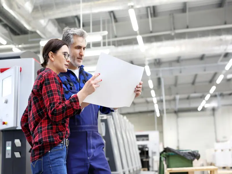 A young woman and an older man review a printed document in a pressroom. The woman is wearing a red plaid shirt and jeans and the man is in blue overalls with a blue flannel shirt.