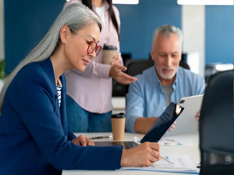 A woman with grey hair and glasses reviews paperwork with a younger person looking on.