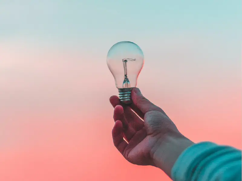An extended arm holding a light bulb against a hazy red and blue sky.