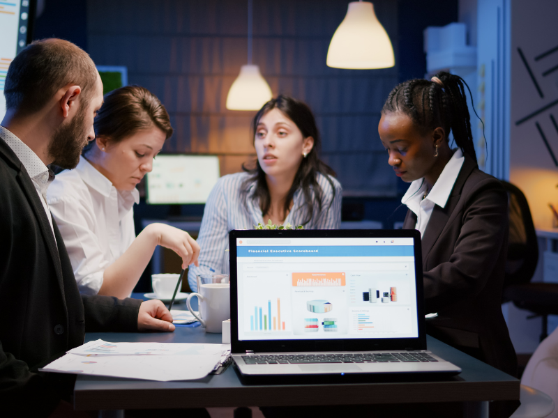 A diverse group of business professionals working together at a table, with financial data displayed on a laptop.
