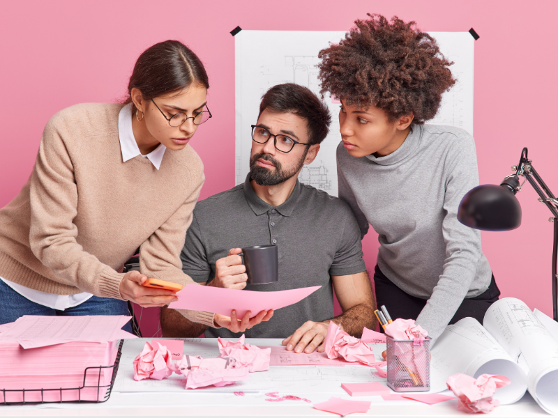 Three colleagues reviewing design plans at a desk with crumpled papers against a pink background.