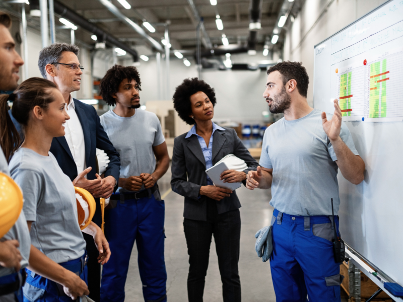 A group of workers and managers in a factory discussing production efficiency in front of a whiteboard with data charts.