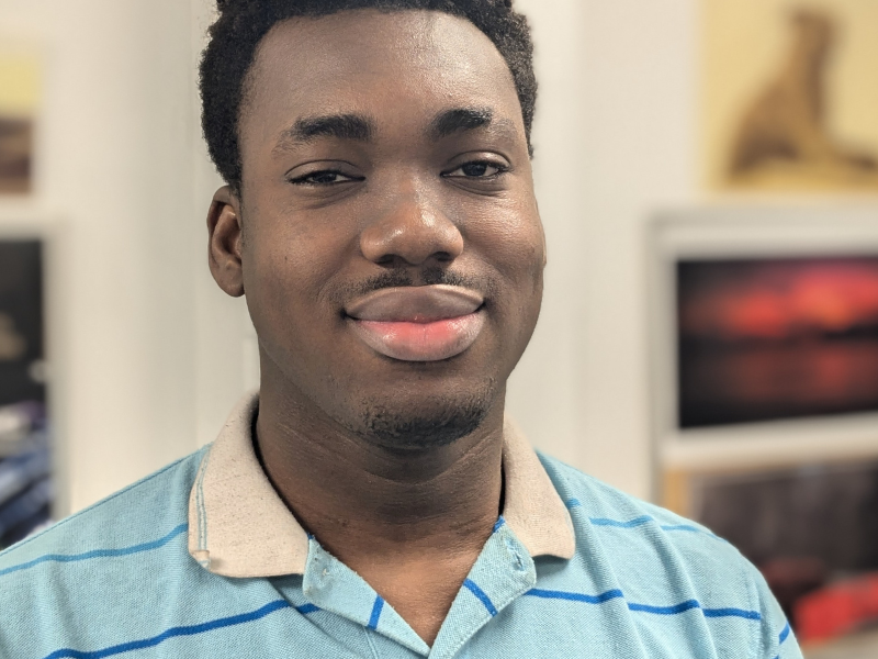 A young man wearing a striped shirt, smiling confidently while standing in a room with colorful artwork in the background.