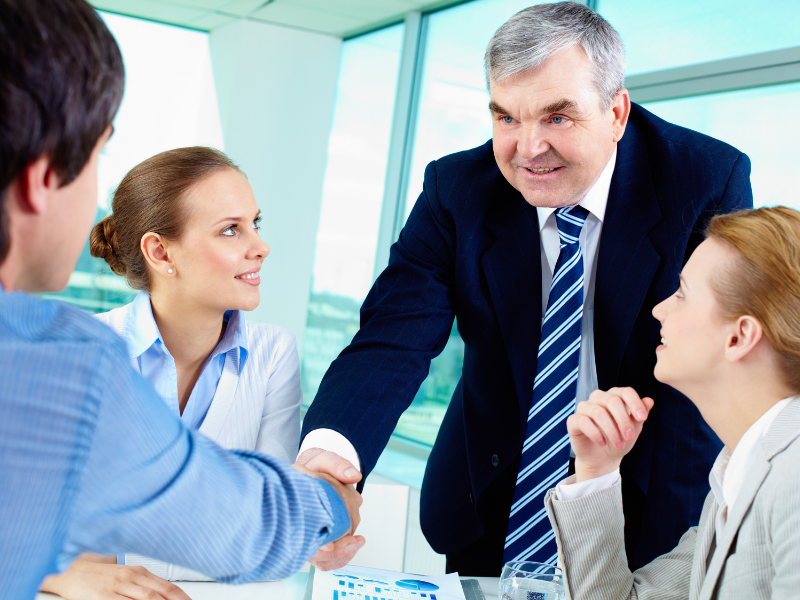 A business professional shaking hands with a colleague across a conference table, surrounded by team members in a modern office.