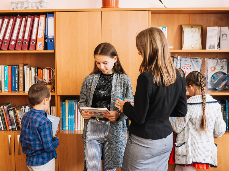 Students and a teacher browsing books in a library with shelves filled with books and binders in the background.