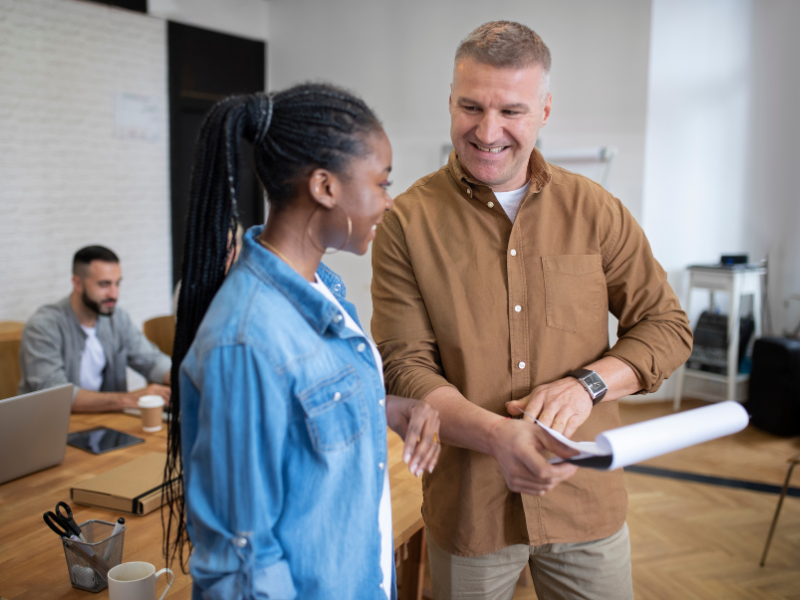 A smiling manager welcomes a new team member in a modern office, holding a document while engaging in conversation.