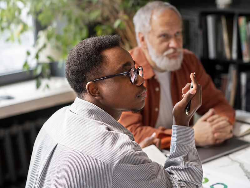A young man listens intently during a mentoring session, seated beside an older advisor in a print industry office setting.