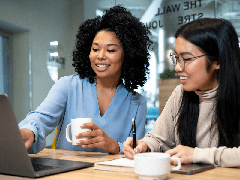 Two women engaged in a mentoring session, smiling while reviewing information on a laptop in a modern office setting.