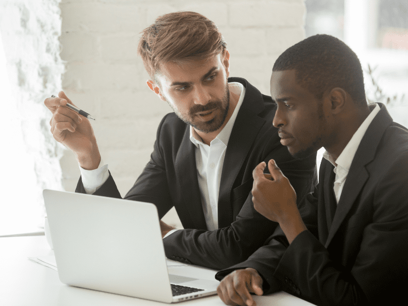 Two business professionals in suits discuss strategy while working on a laptop in a bright office, illustrating executive coaching and leadership development.