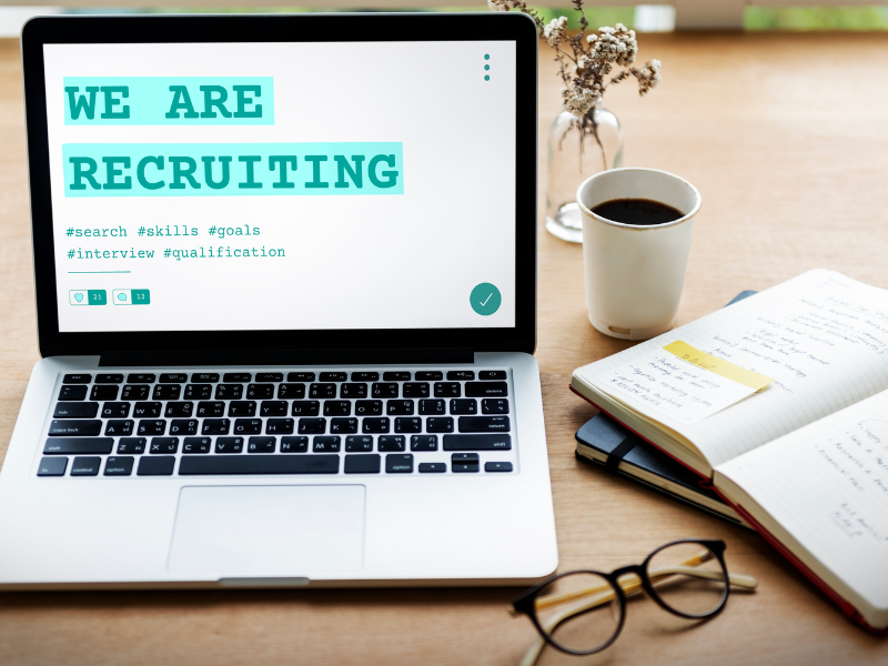 Laptop displaying “We Are Recruiting” beside a notebook and coffee, reflecting a proactive hiring search for complex roles.