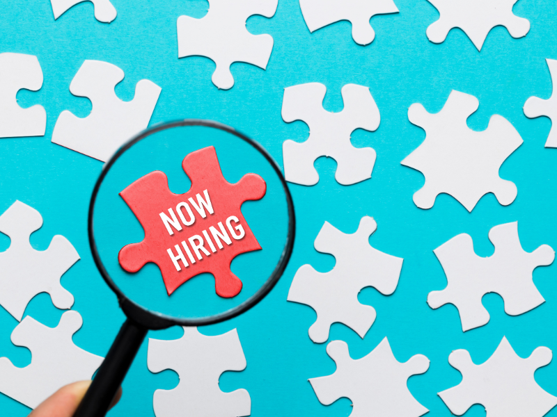 Magnifying glass highlights a red “Now Hiring” puzzle piece among white pieces, reflecting recruiting and career path gaps.