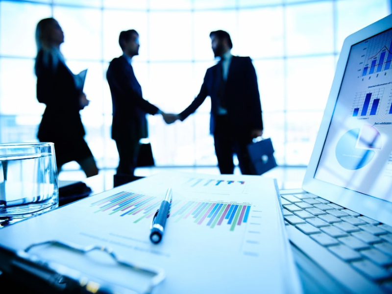Executives review financial charts as two leaders shake hands, reflecting M&A valuation and capability gap analysis.
