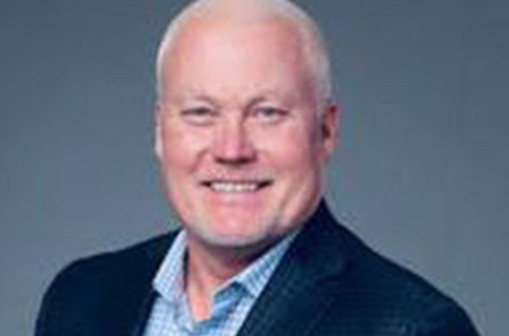A headshot of management consulting expert, Darren Loken. Darren is male and wearing a dark navy suit with light blue button up shirt. He is in front of a grey background.