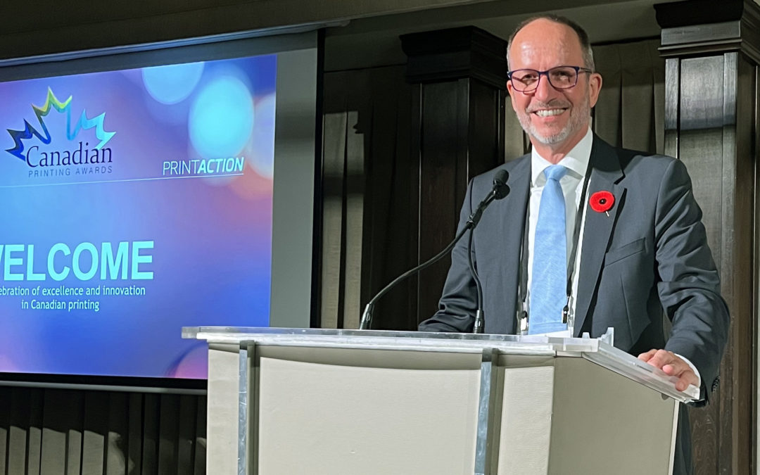 Ken Freek standing at the podium at the Canadian Printing Awards gala. Ken is male, is smiling and is wearing a suit with a blue tie and a poppy.