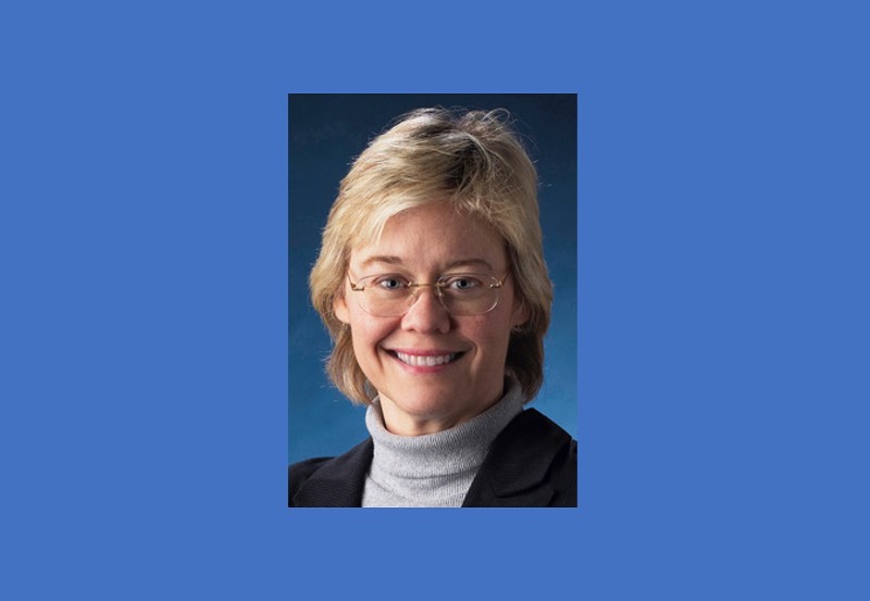 A headshot of Nicky Milner. Nicky is smiling, has short blonde hair, wears glasses and a grey turtleneck. She is against a dark blue backdrop.