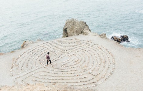 A man journeys through a maze made out of stones on a sea-side cliff.