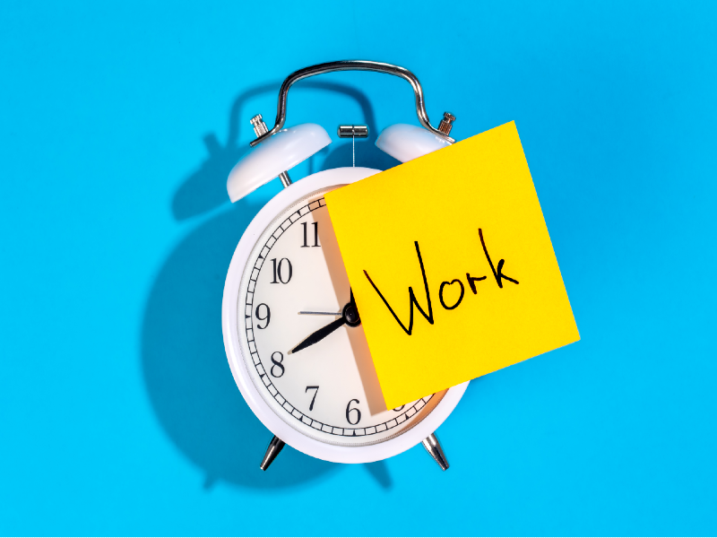 A white alarm clock with a yellow post it note with "work" written on it against a turquoise background
