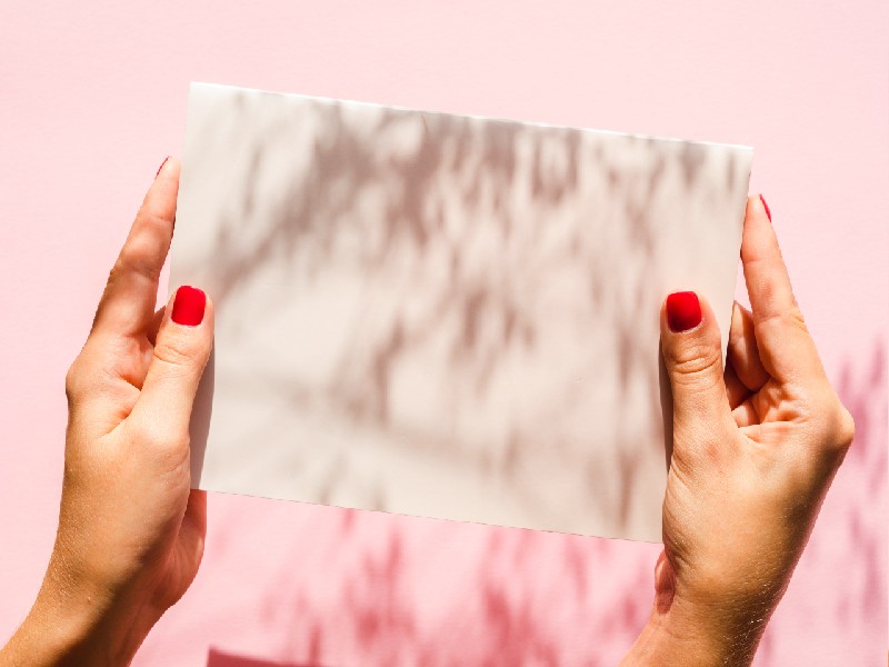 Hands of a young woman with red painted nails hold either side of a white piece of paper against a pale pink background.