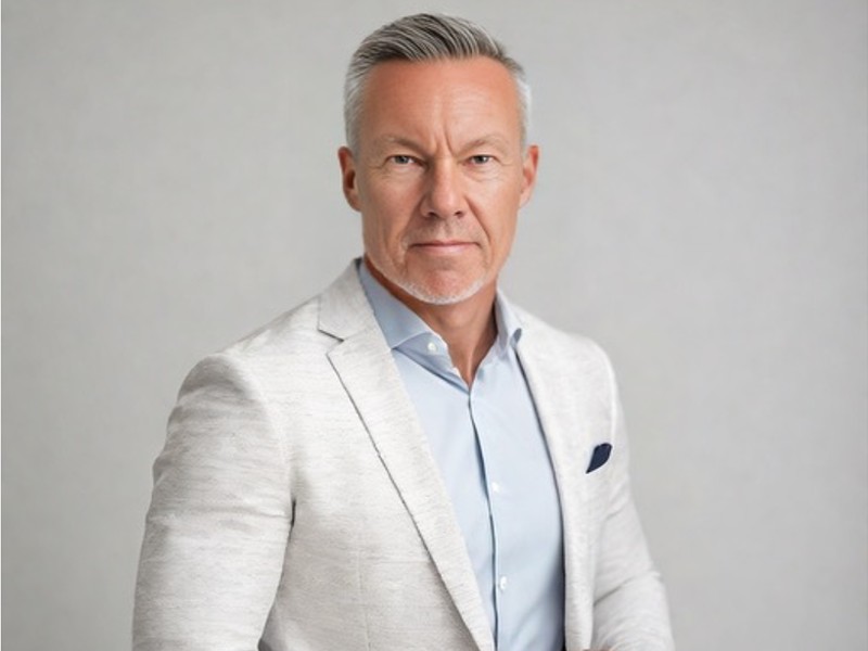 A man with grey hair wearing a white blazer and blue button up shirt.