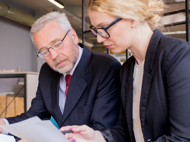 Two professionals, a mature man in a suit and a younger woman in business attire, reviewing a document together in a modern office setting.