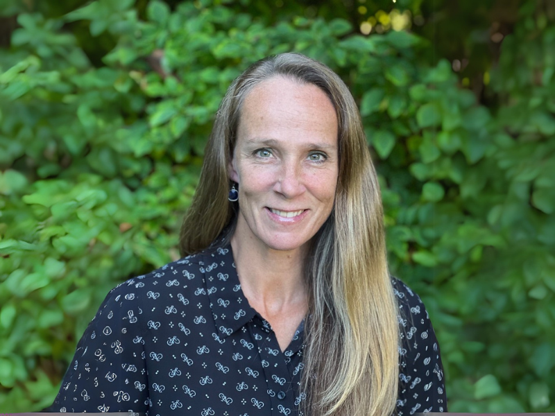 Portrait of Julie Anne McLaren smiling, standing in front of green foliage.