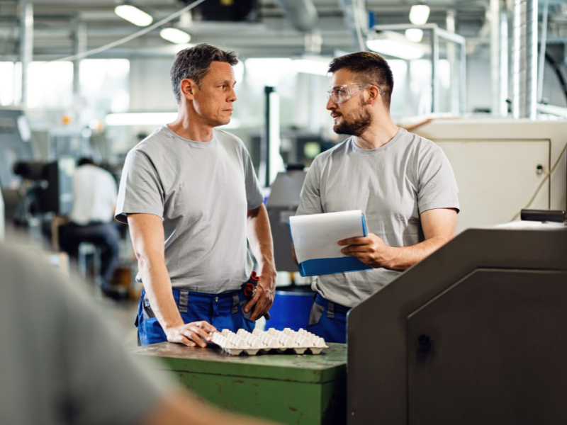 Two print shop employees discussing a task on the production floor, with machinery and tools in the background.