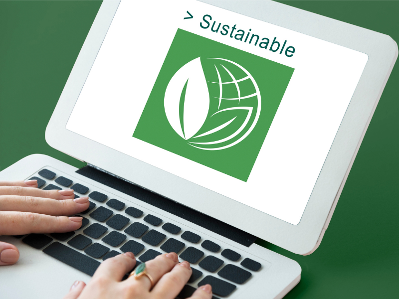 A laptop screen displaying a green sustainability icon with a globe and leaf, with hands typing on the keyboard.