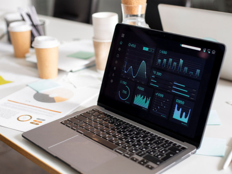 A laptop displaying data analytics dashboards with various charts and graphs, placed on a desk with printed reports and coffee cups.