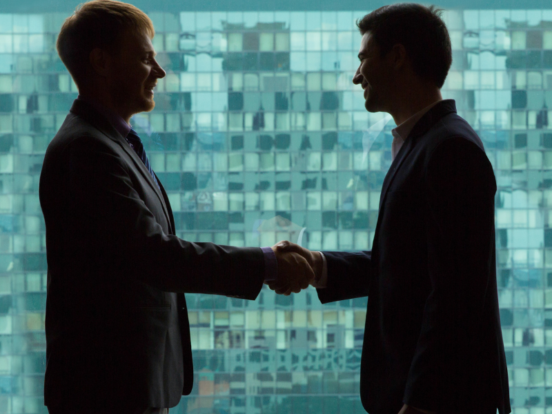 Two business professionals shaking hands in front of a modern glass building, symbolizing a successful negotiation and mutual agreement.