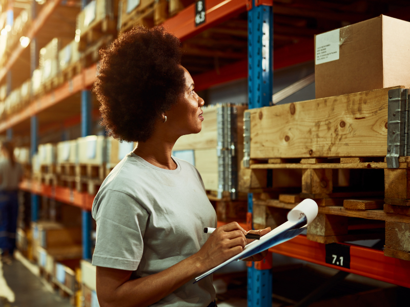 A warehouse employee holding a clipboard, inspecting inventory on wooden pallets in a well-organized storage area.